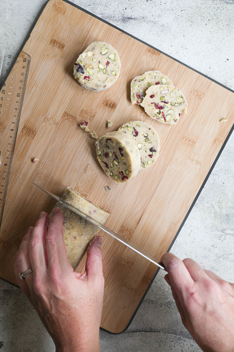 2 hands slicing the cranberry pistachio shortbread cookies with a knife on a wooden chopping board.
