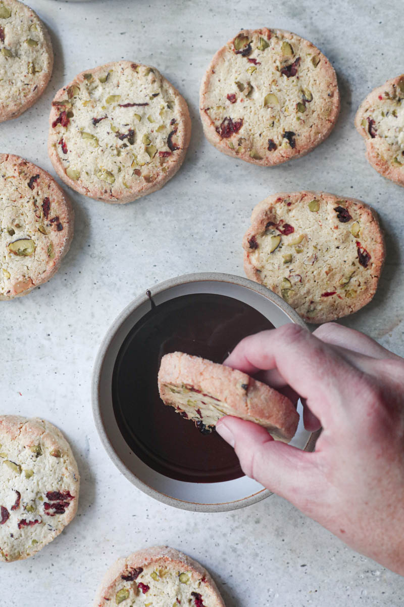 One hand dipping a cranberry pistachio shortbread cookie in a bowl full of melted chocolate with other cookies around.