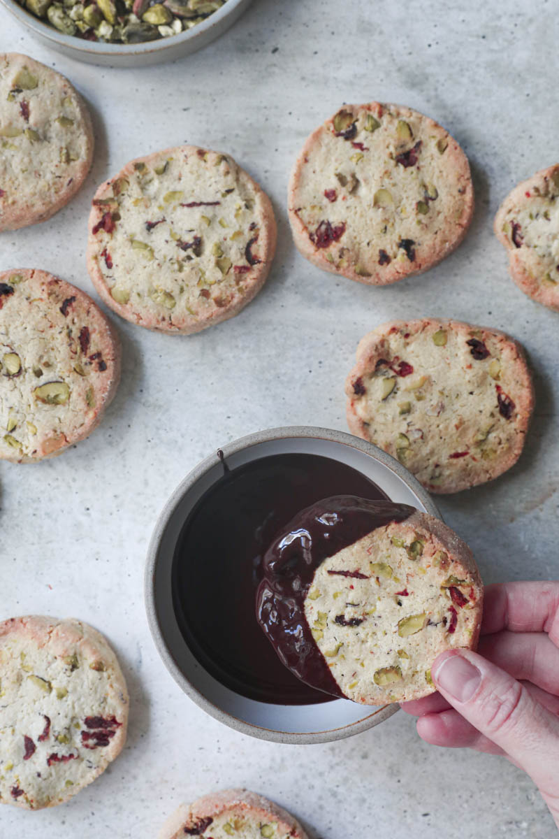 One hand dipping a cranberry pistachio shortbread cookie in a bowl full of melted chocolate with other cookies around.