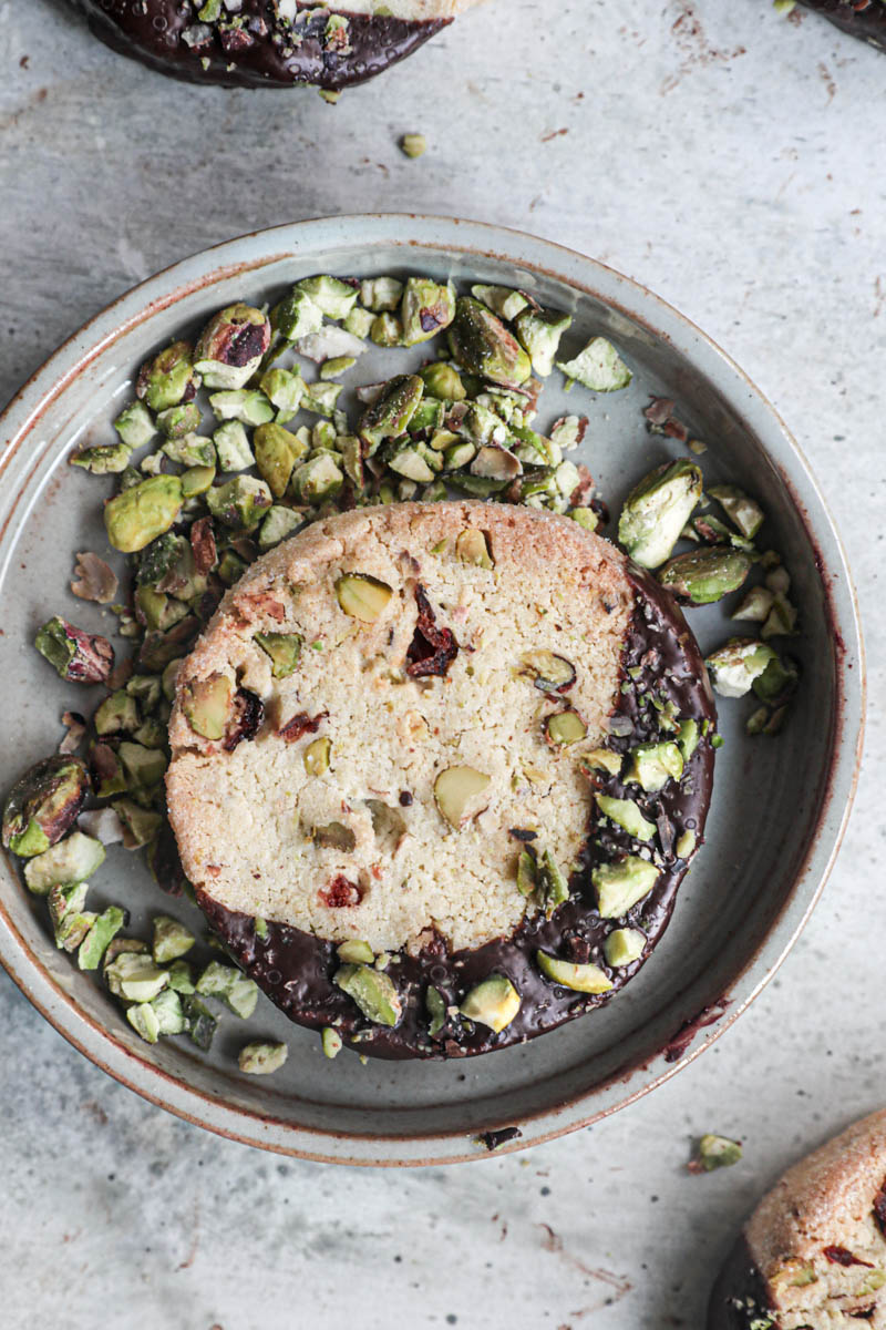 One shortbread cookie dipped in chocolate drizzled with crushed pistachios on a green plate with pistachios all around.