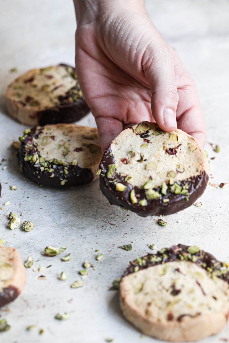 One hand holding one shortbread cookie dipped in chocolate drizzled with crushed pistachios with other cookies around.