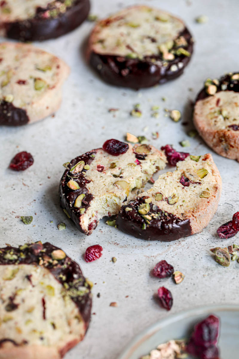 One cranberry pistachio chocolate dipped shortbread pistachio cookie with others on the side and some dried cranberries around.