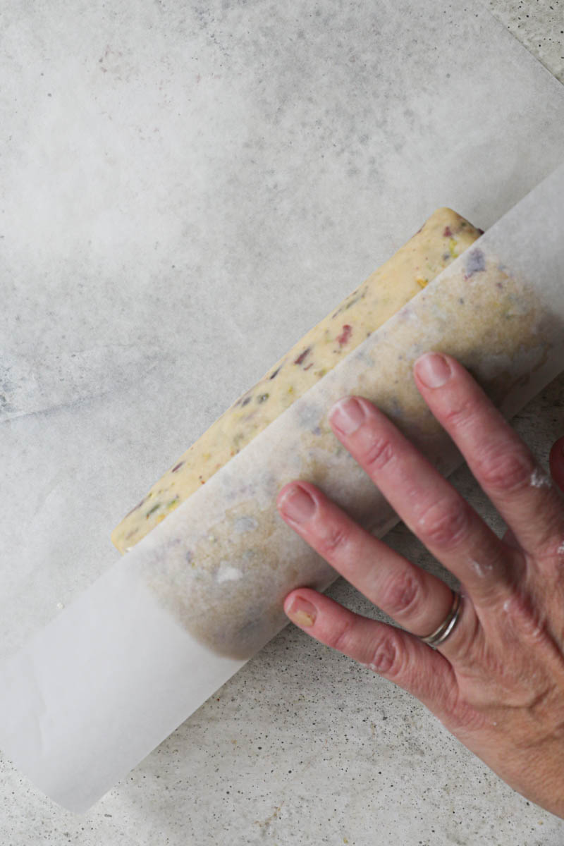 2 hands covering the cranberry pistachio shortbread cookie dough shaped as a log with parchment paper.