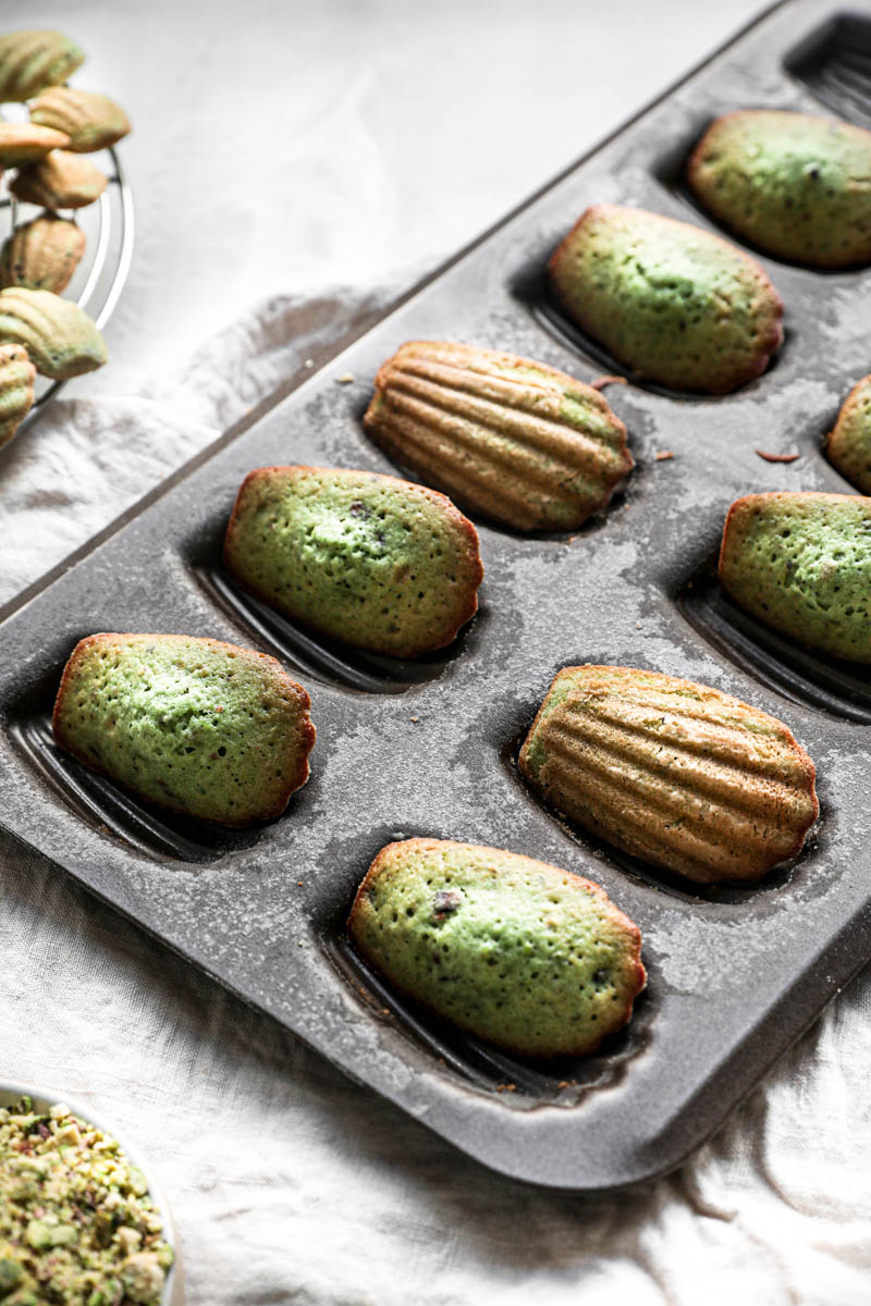 The baked pistachio madeleines inside the madeleine pan, turned 90 degrees with other madeleines on wire rack in the left top corner.