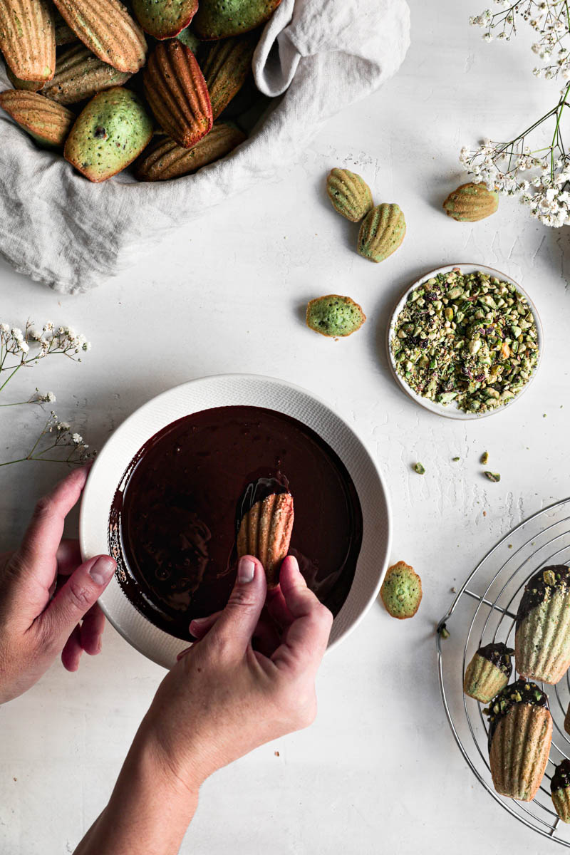 One hand dipping a madeleine inside a bowl of chocolate glaze with a wire rack filled with baked madeleines in the top left corner and plate full of grounded pistachio on the side.