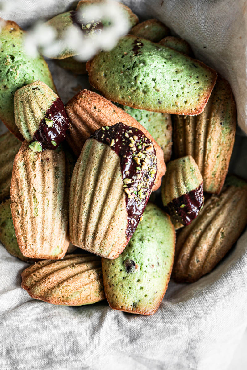 A basket covered in a beige linen holding the pistachio madeleines covered in chocolate glaze and pistachios as seen from above with some flowers blurry in the corner.