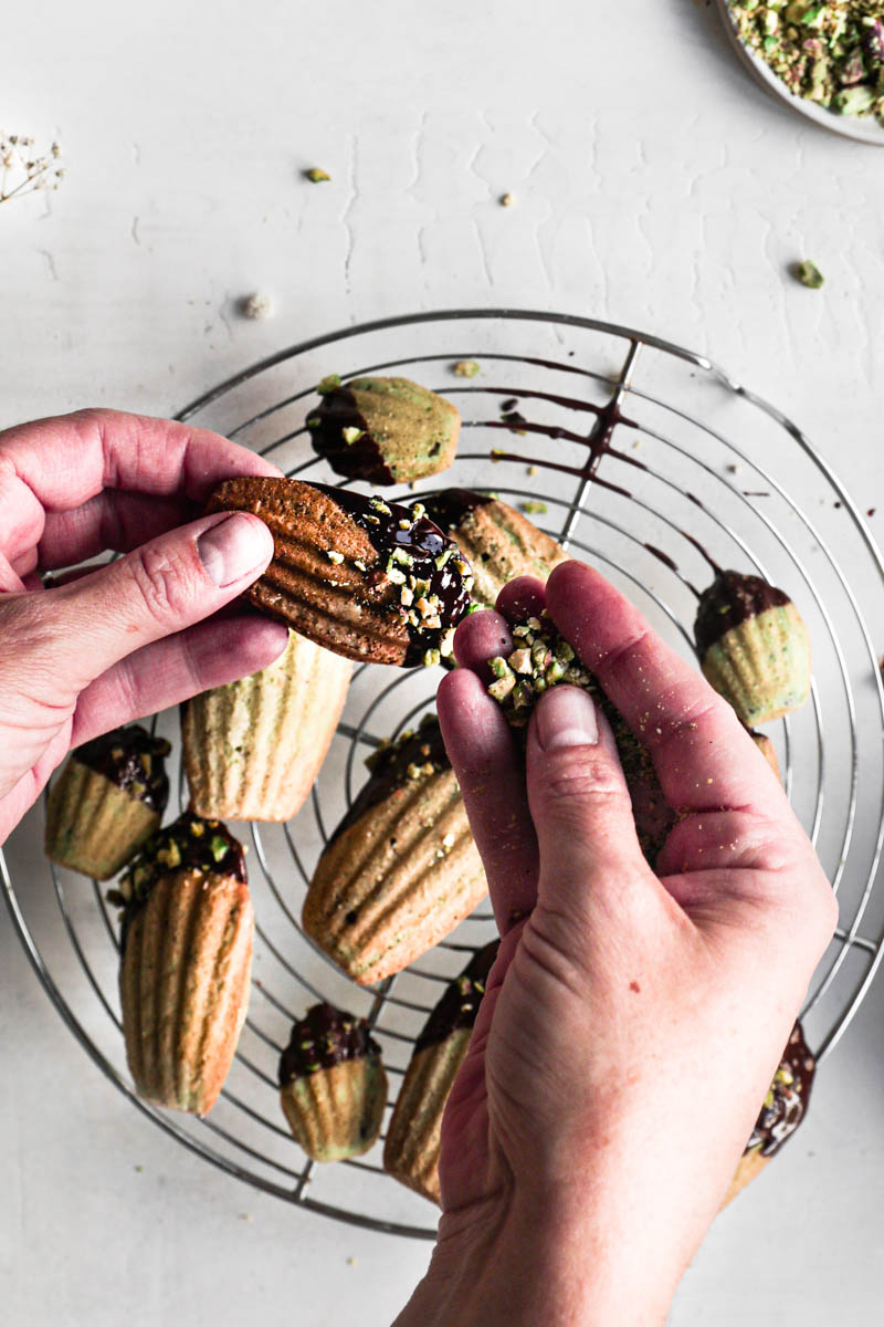 One hand holding a glazed pistachio madeleine and the other hand sprinkling it with some grounded pistachios with a blurry wire rack filled with more madeleines right underneath.