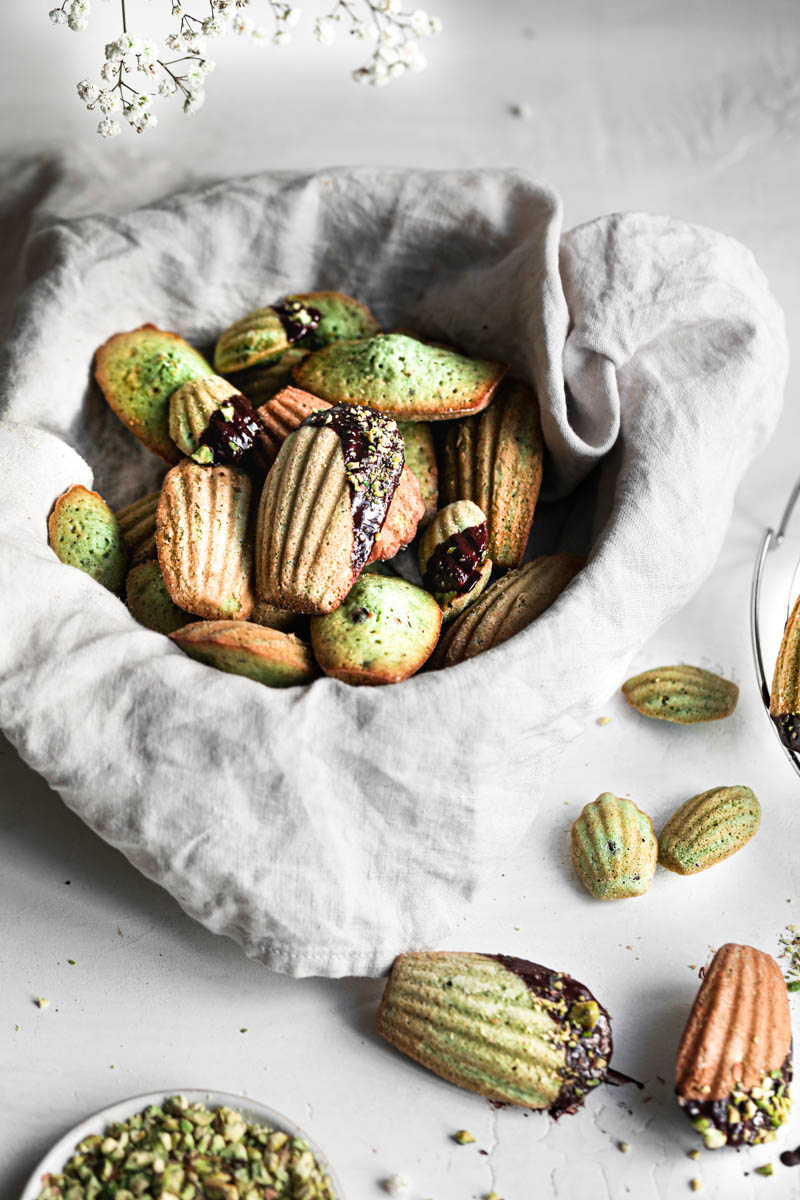 A basket covered in a beige linen holding the pistachio madeleines covered in chocolate glaze and pistachios and some madeleines surrounding the basket.