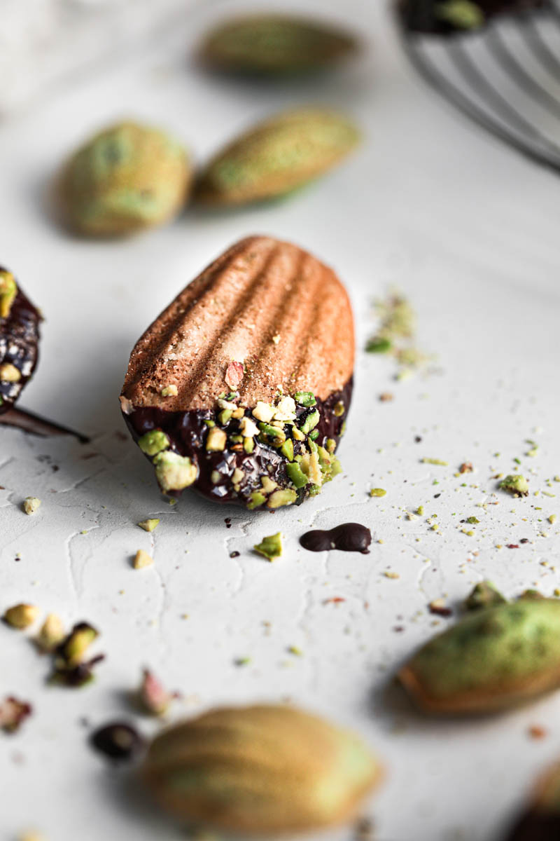 One pistachio French madeleine covered in chocolate glaze in focus at the center of the frame with other small madeleines around it.