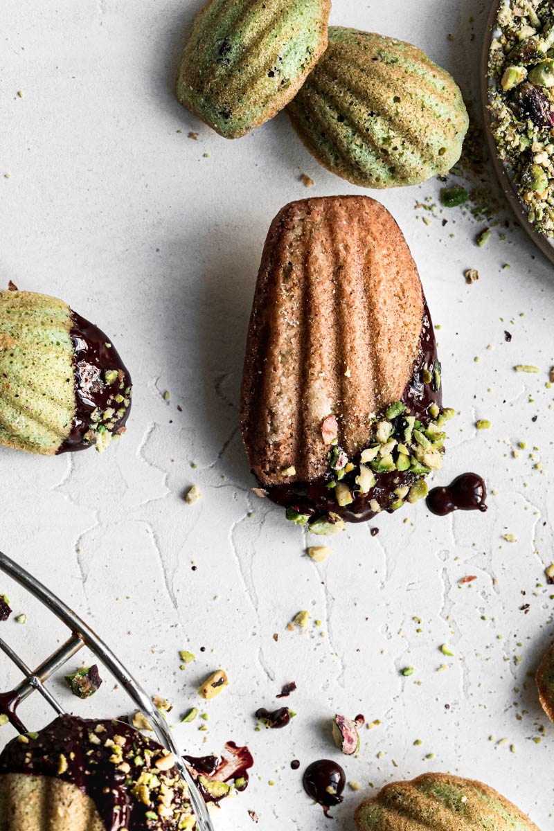 One pistachio French madeleine covered in chocolate glaze in focus at the center of the frame with other small madeleines around it as seen from above.