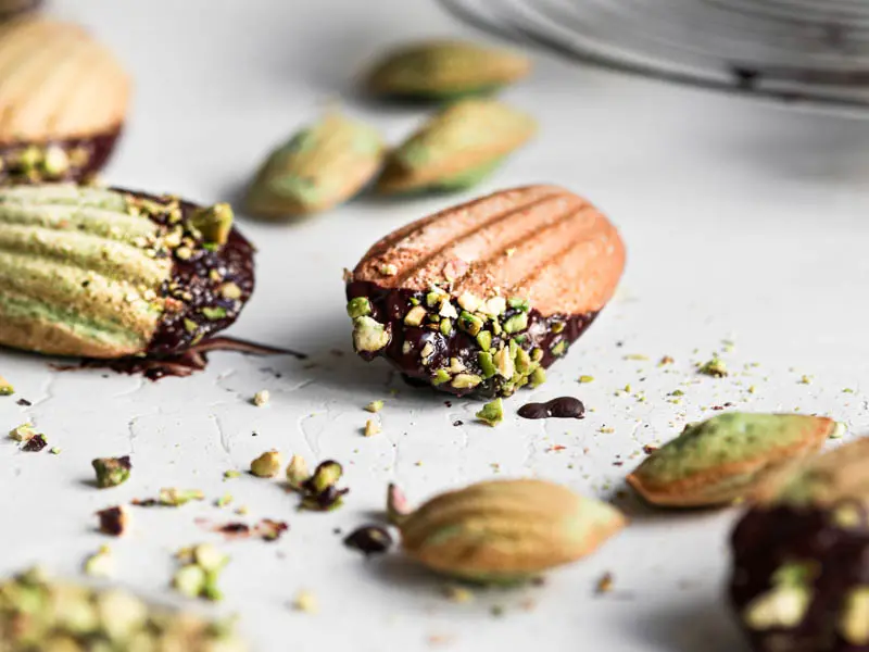 One pistachio French madeleine covered in chocolate glaze in focus at the center of the frame with other small madeleines around it.