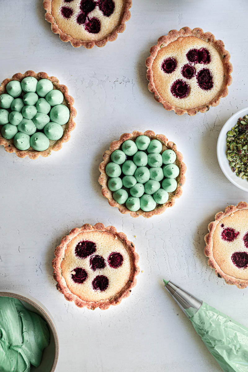 Pistachio raspberry white chocolate tartlets assembly: some tartlets topped with the ganache, others not yet, and a piping bag on the side.