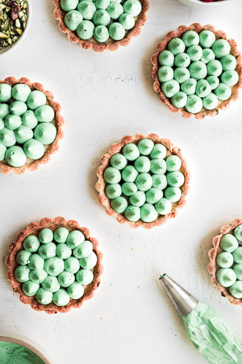 Pistachio raspberry white chocolate tartlets assembly: all the tartlets topped with white chocolate pistachio ganache kisses and a piping bag on the side.