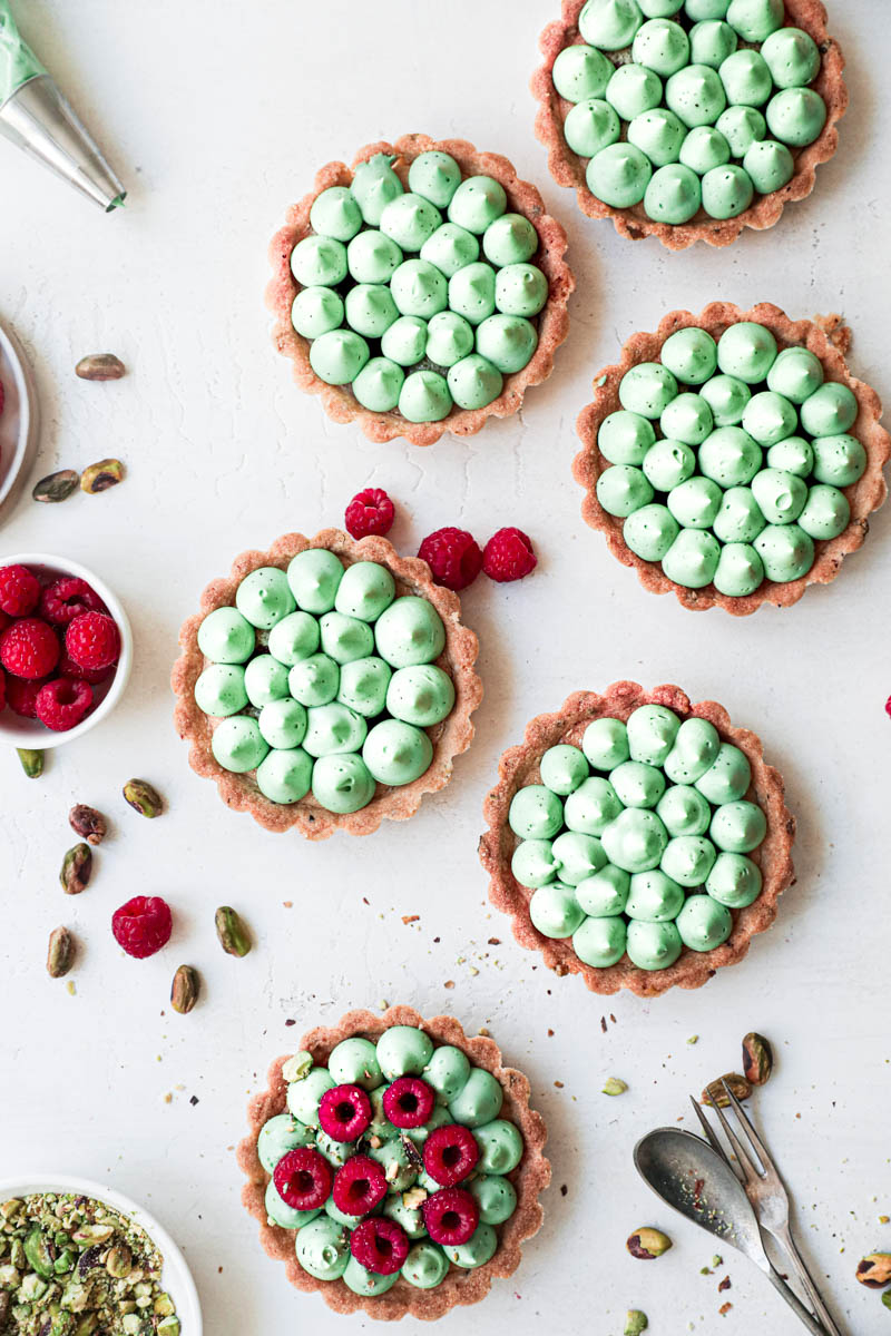 Pistachio raspberry white chocolate tartlets assembly: one tartlet topped with fresh raspberries and the other five only with ganache kisses.