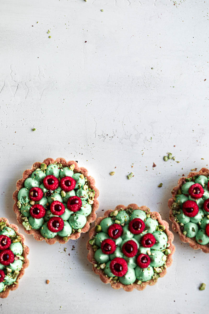 4 pistachio raspberry white chocolate tartlets assembled topped with fresh raspberries places in a zigzag at the bottom of the frame.
