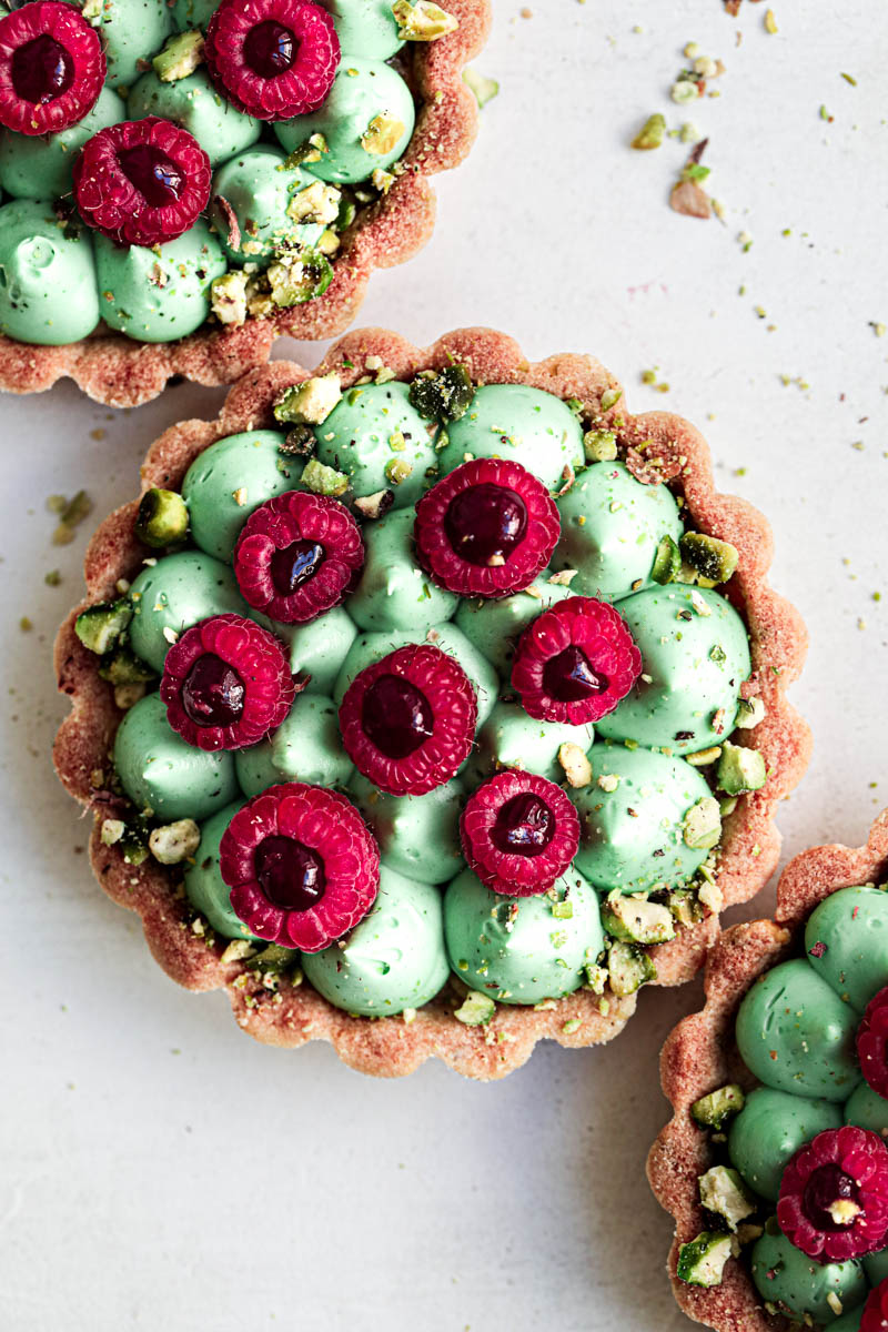 A closeup shot of 3 pistachio raspberry white chocolate tartlets assembled topped with fresh raspberries.