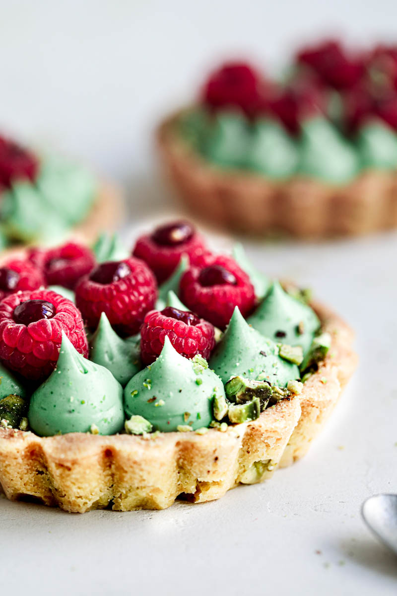 Closeup of 1 pistachio raspberry white chocolate tartlet with two others blurry in the back.