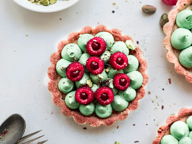 Overhead shot of 1 pistachio raspberry white chocolate tartlet with the edges of 2 others showing on the sides.