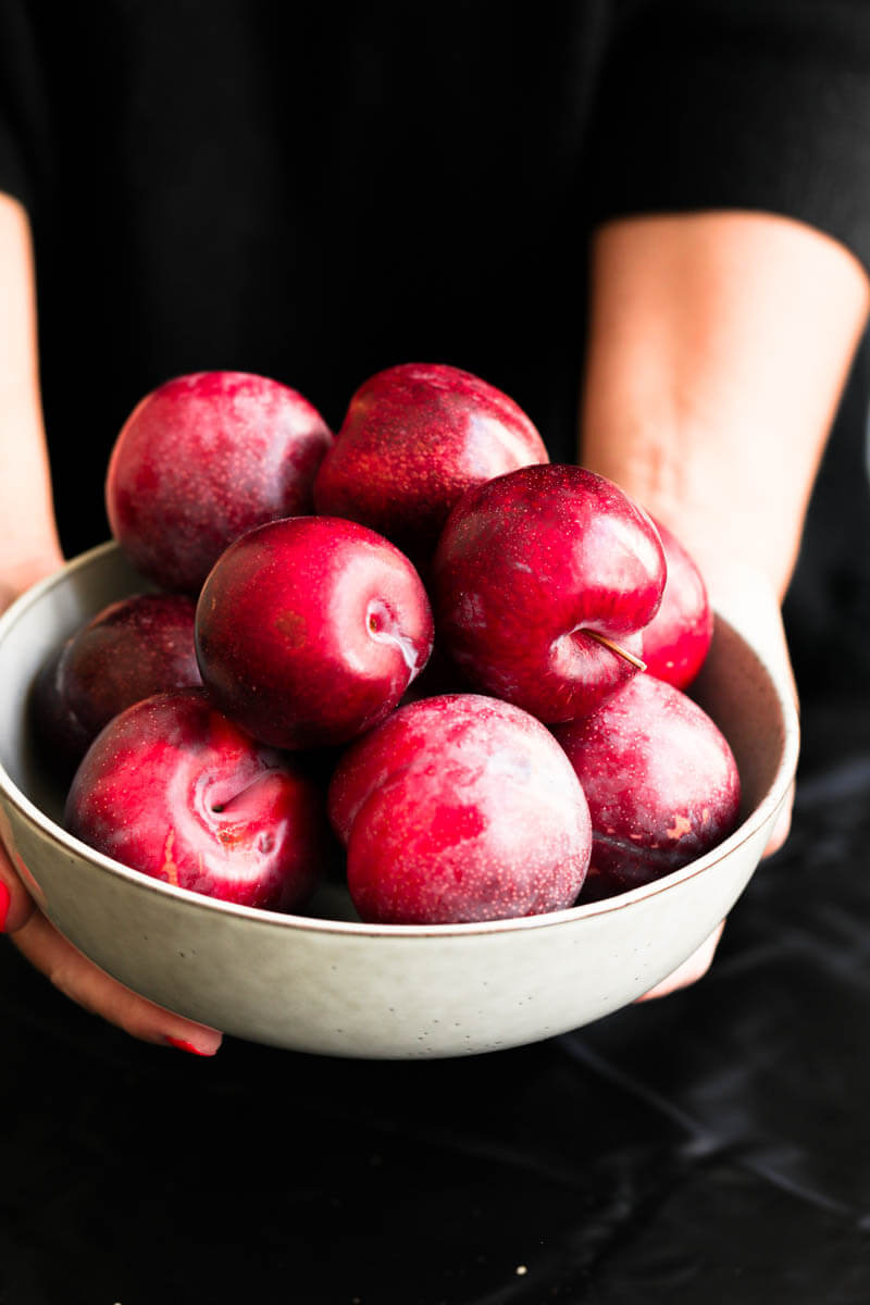 90° shot of 2 hands holding a bowl with fresh plums