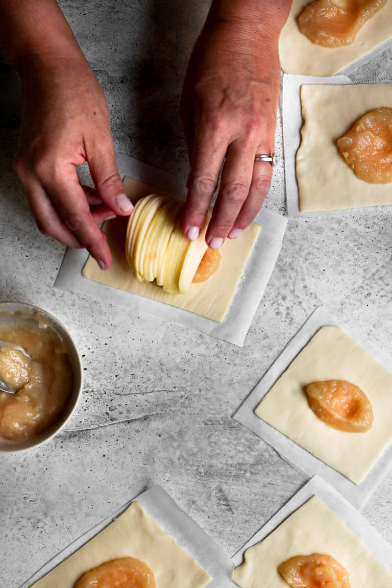 Overhead shot of 2 hands placing half a sliced apple over the puff pastry dough