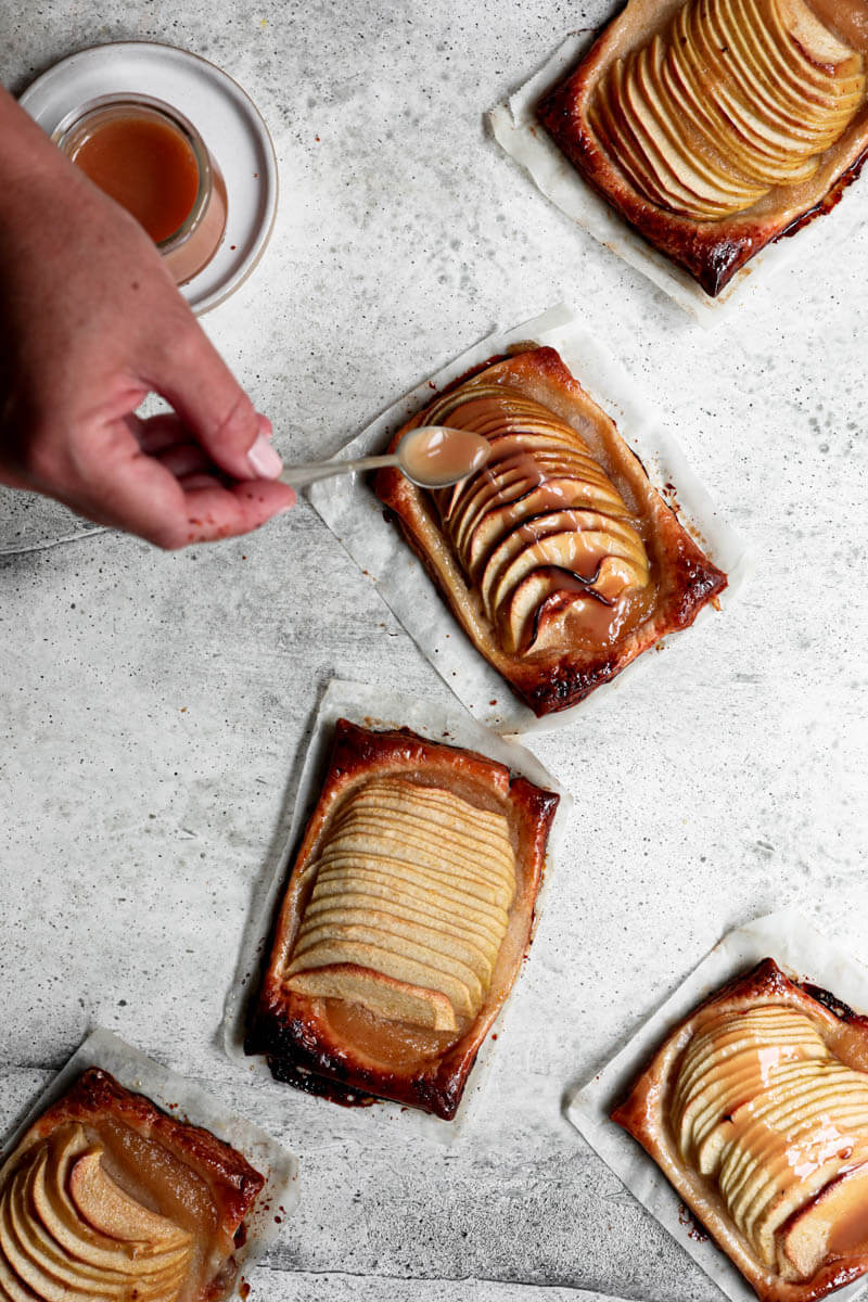 Overhead shot of the puff pastry apple tarts with a pot of salted caramel in the top left corner with a hand drizzling salted caramel over one tart