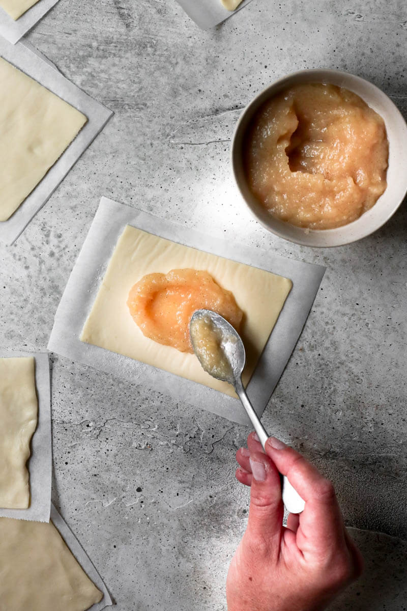 Overhead shot of one hand filling a puff pastry rectangle with apple compote