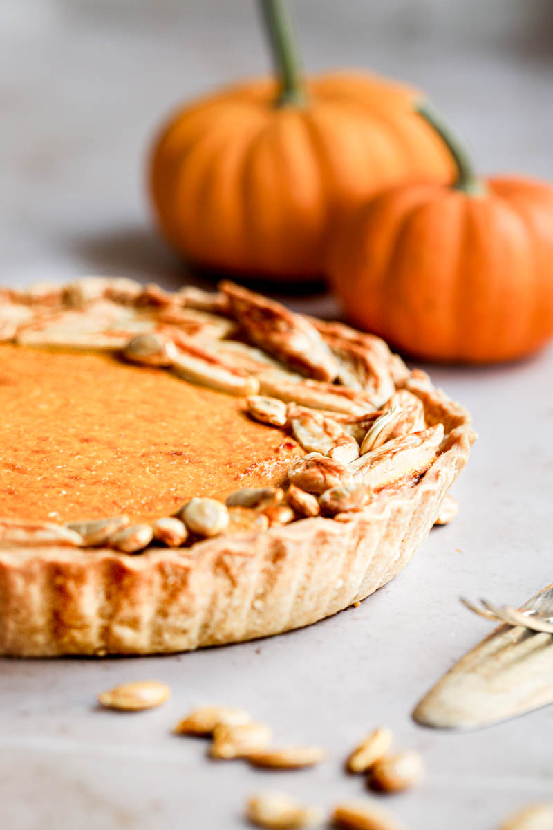 Three quarters of the pumpkin pie seen from the side with 2 pumpkins seen behind and some pumpkins seeds on the side of the frame.