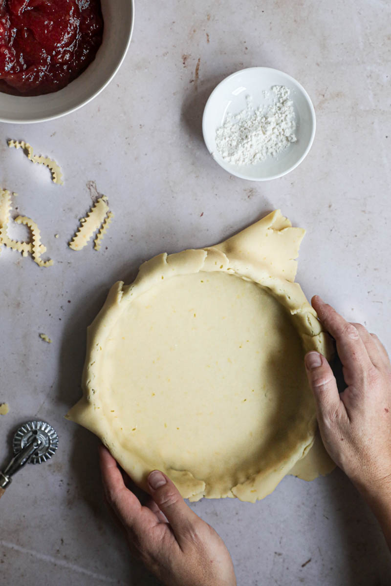 Two hands holding the tart in lined with the quince pie dough with a bowl with quince pie filling in the top corner of the frame.