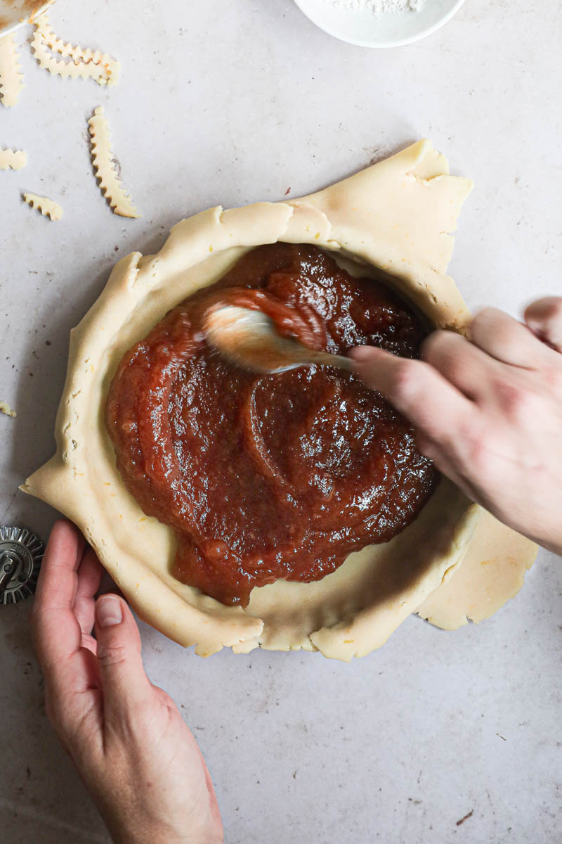 One hand spreading the quince pie filling over the tart base using a spoon.