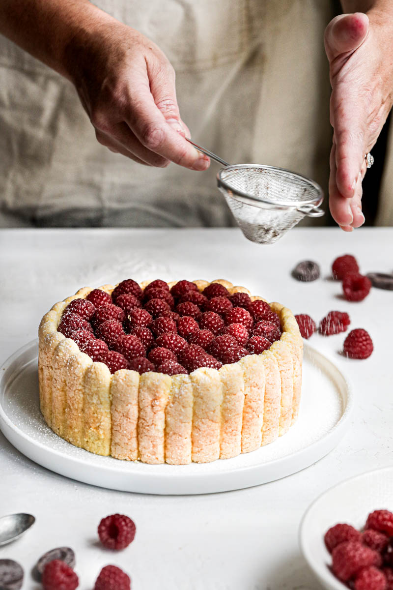 Two hands sifting powdered sugar over the fresh raspberry chocolate charlotte using a small sifter.