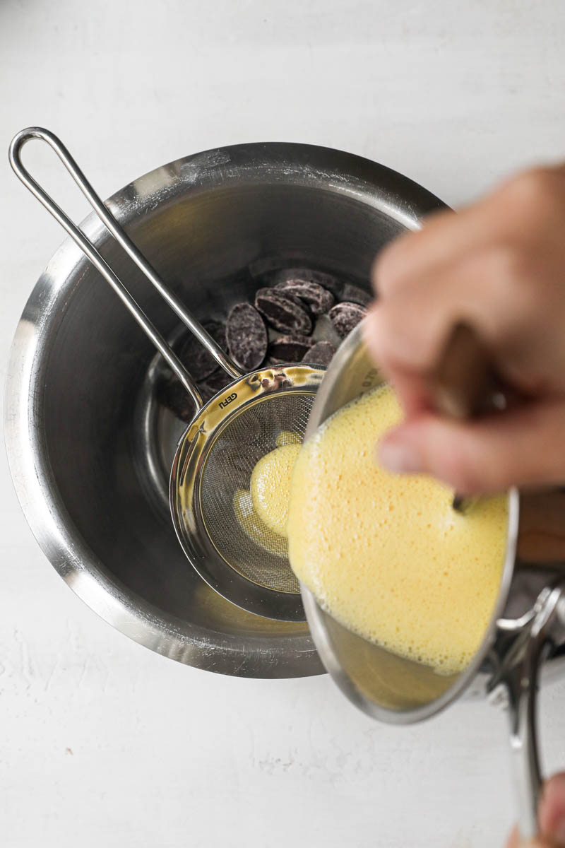 A bowl with chocolate with a strainer placed over it a hand pouring the custard over it.