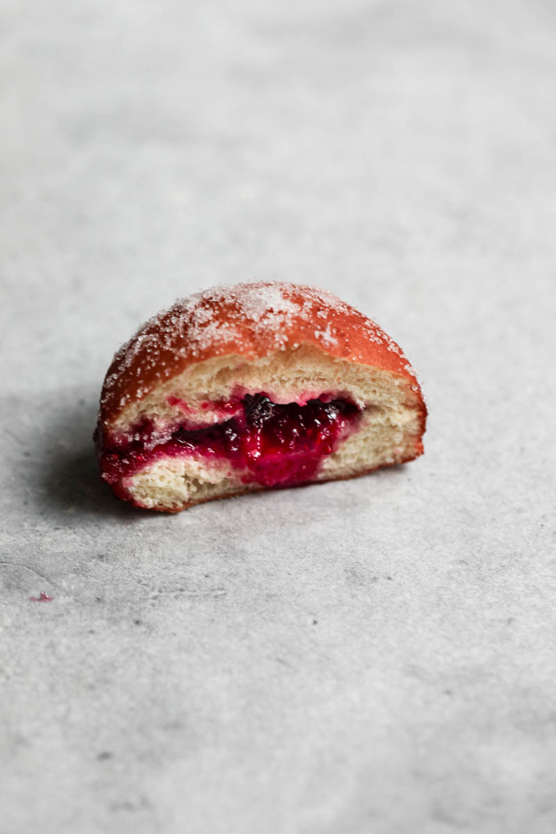 A sliced filled raspberry jam brioche donut and a corner of the other half in the bottom right angle.