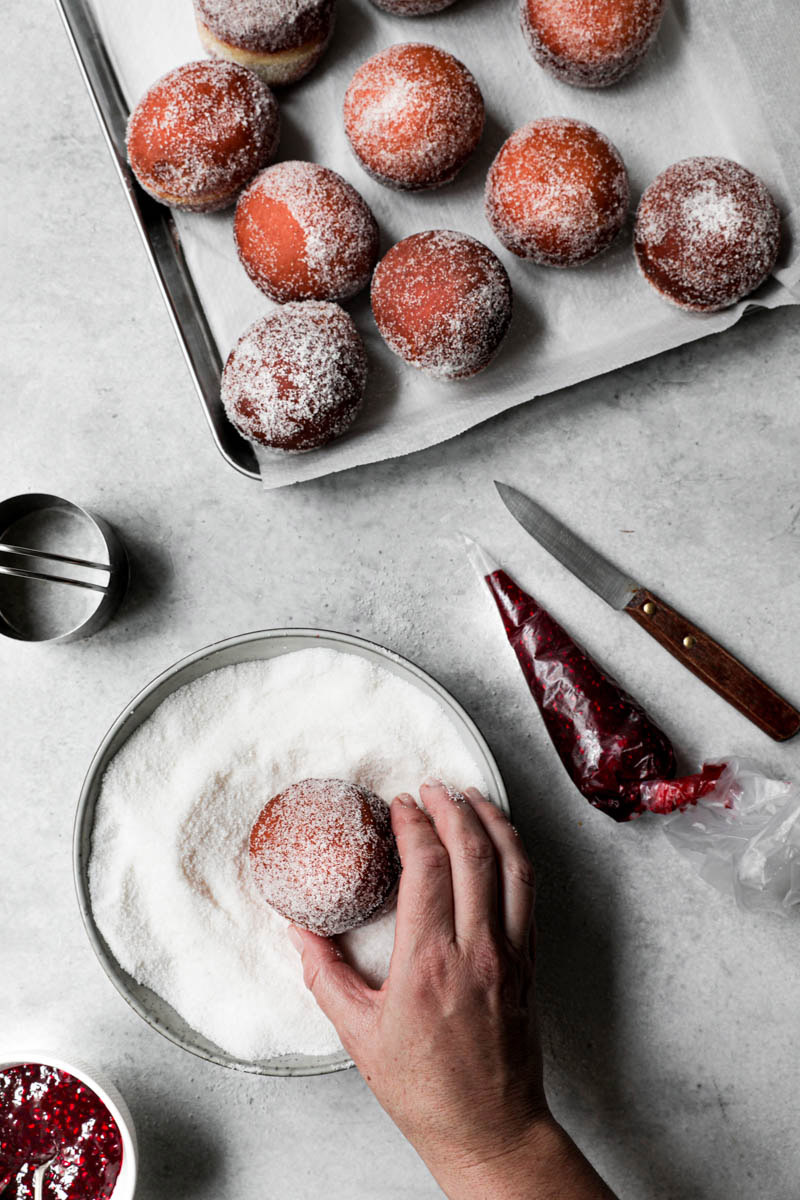 A hand rolling a deep-fried donut in sugar. A baking tray filled with sugar coated donuts in the top corner of the frame.