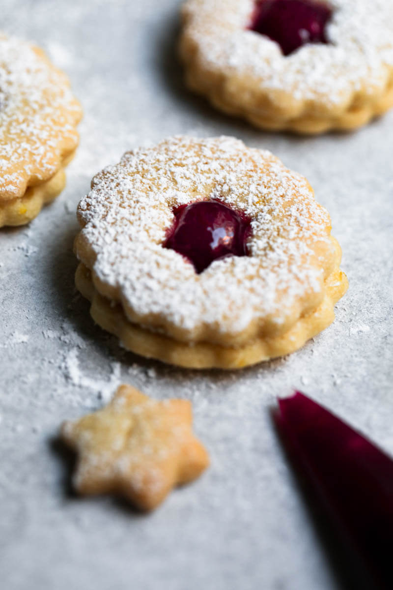 One linzer cookie filled with raspberry jam, with other blurry behind and a piping bag in the bottom right corner