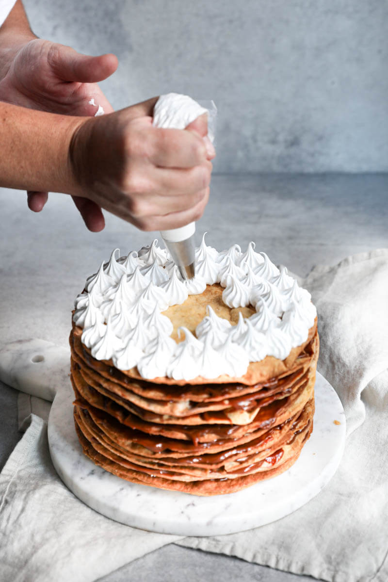 2 hands piping swiss meringue frosting on the dulce de leche cake
