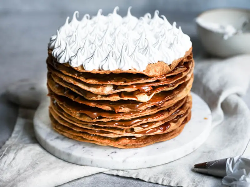 Whole rogel cake frosted with swiss meringue with a bowl behind it out of focus