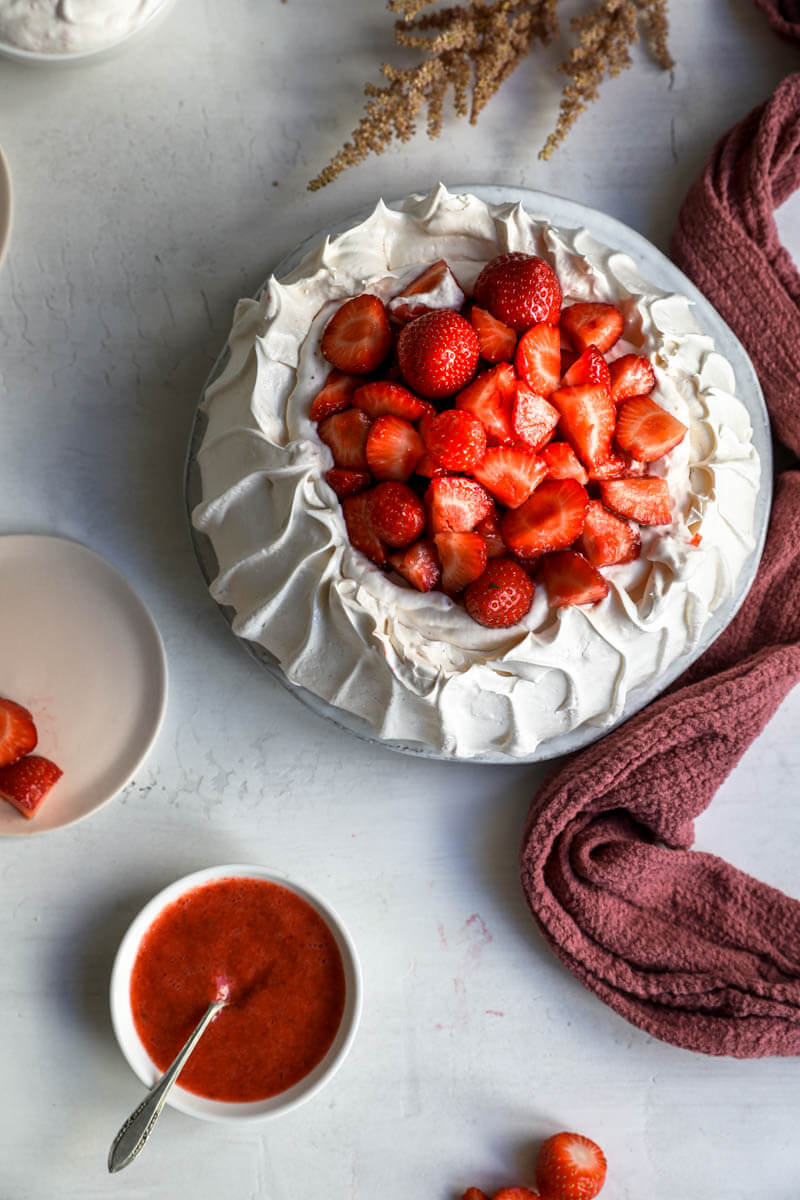 Overhead shot of the meringue base filled with strawberry Chantilly cream and topped with fresh strawberries surrounded by flowers on the to corner and a bowl filled with strawberry compote