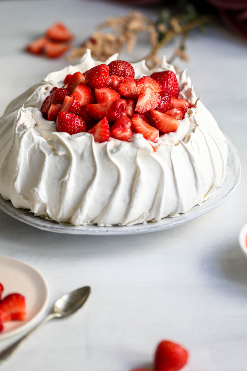 45° shot of the strawberry pavlova served on a white plate with flowers on the background and some fresh strawberries in the front