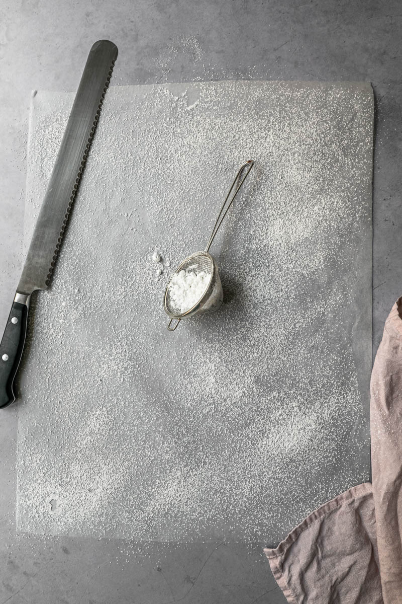 A piece of parchment paper sprinkled with powdered sugar with a strainer and a serrated knife on the side.