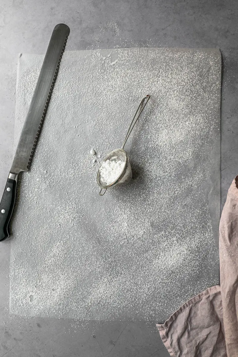 A piece of parchment paper sprinkled with powdered sugar with a strainer and a serrated knife on the side.