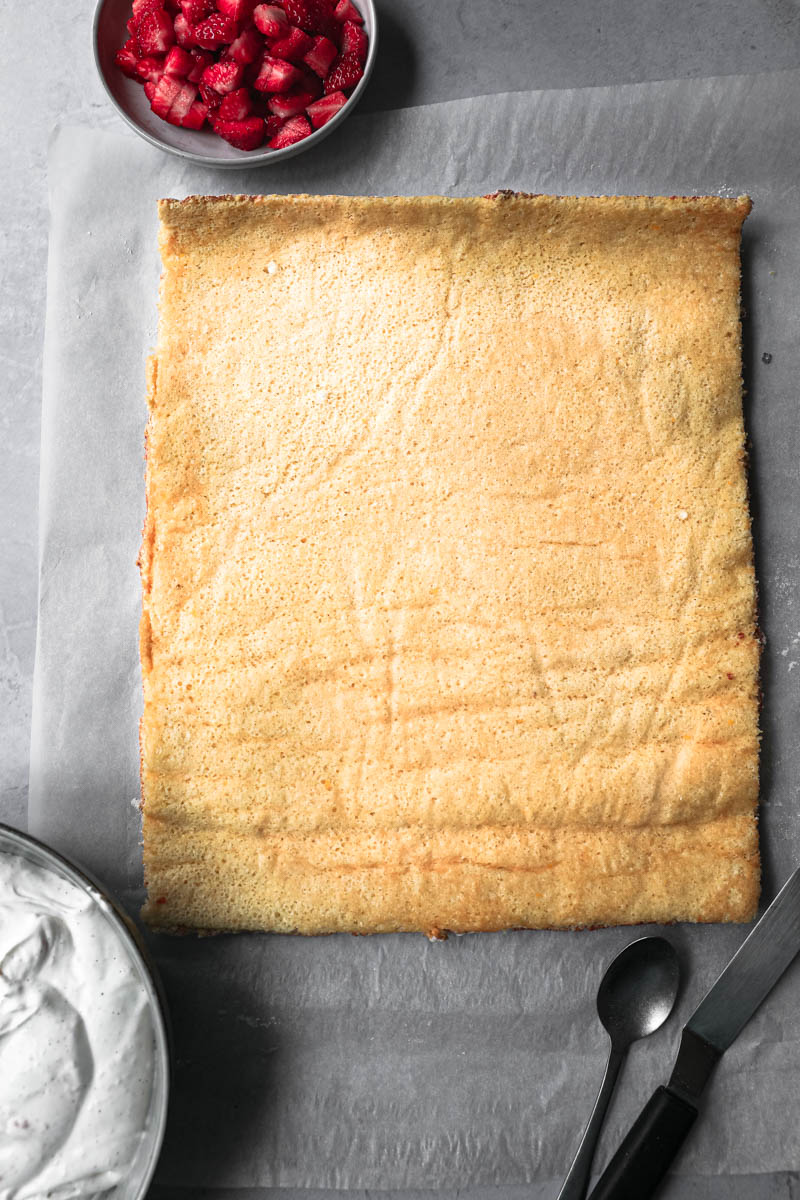 The unrolled cake ready to be filled with a small bowl with diced strawberries on the top lift corner.