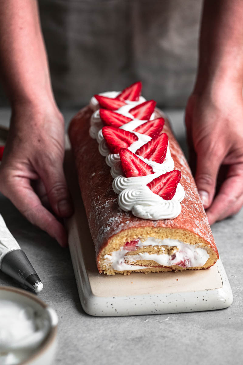 2 hands holding a rectangular plate with a strawberry roll cake on it.