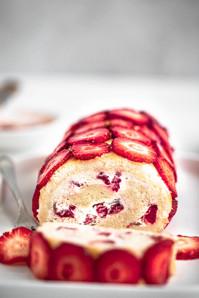 The strawberry roll cake covered with sliced strawberries on a white plate.