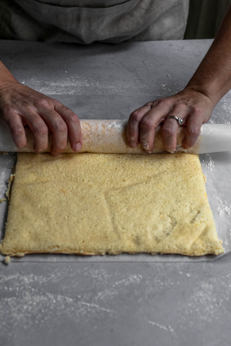 2 hands rolling the roll cake on a piece of parchment paper.