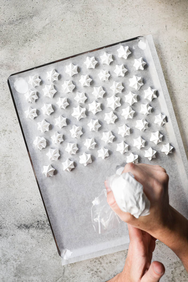 Two hands piping out the meringue kisses on the baking tray lined with parchment paper.