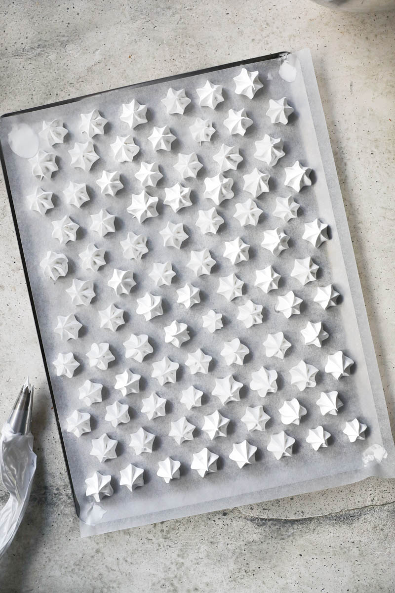 The pipped Swiss meringue kisses on a baking tray ready for the oven with a piping bag on the side as seen from above.