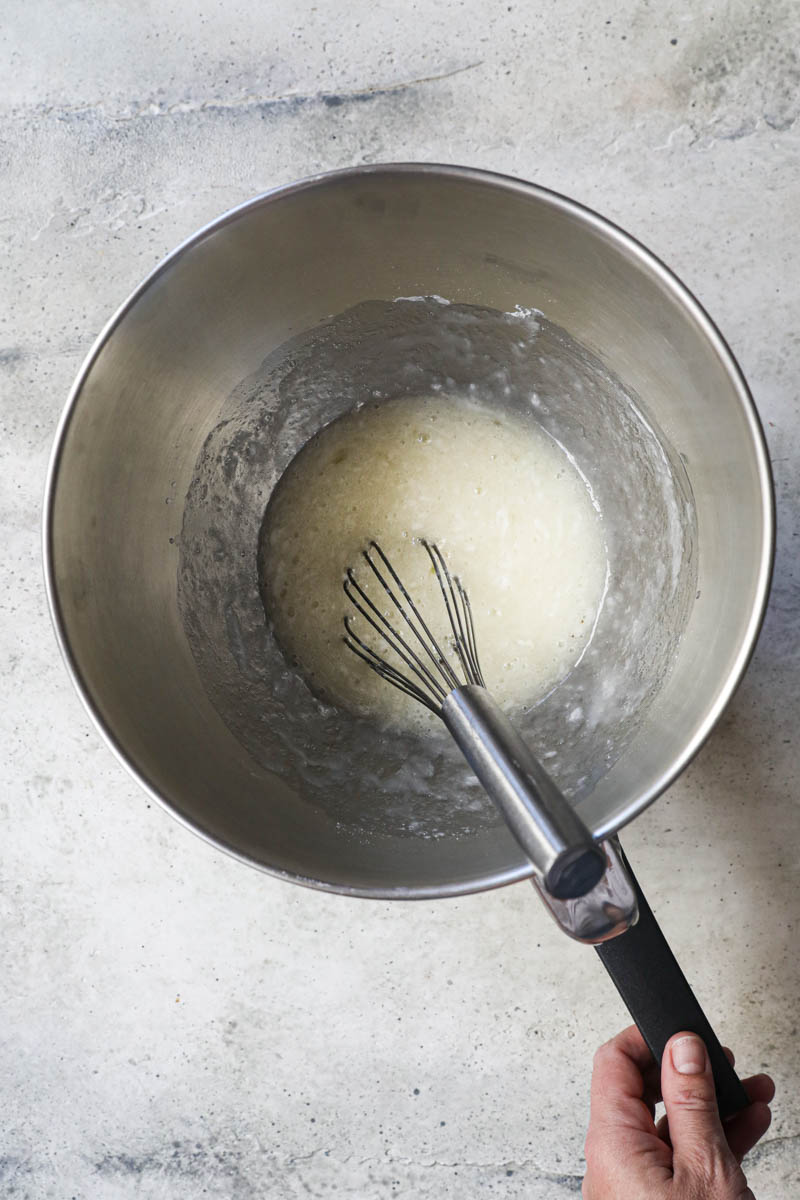 The egg whites and the sugar inside a mixing bowl with a whisk inside over a pan filled with water held by one hand.