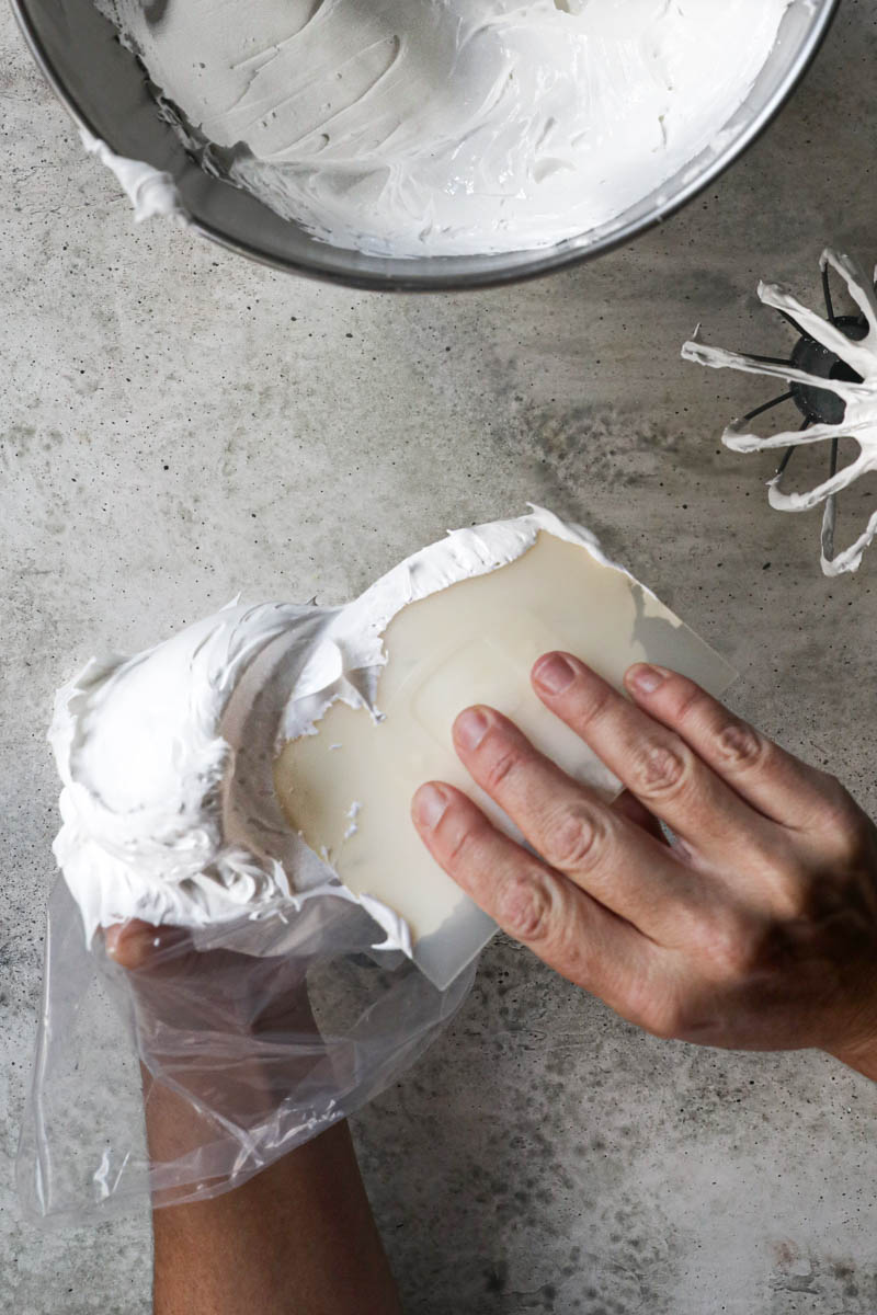 Two hands filling the piping bag with the Swiss meringue using a scraper with a bowl full of meringue on the top of the frame.