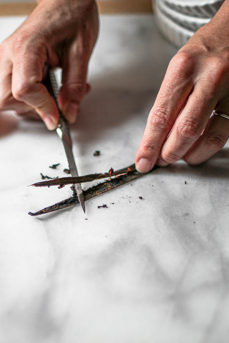 2 hands scraping a vanilla bean using a small paring knife.