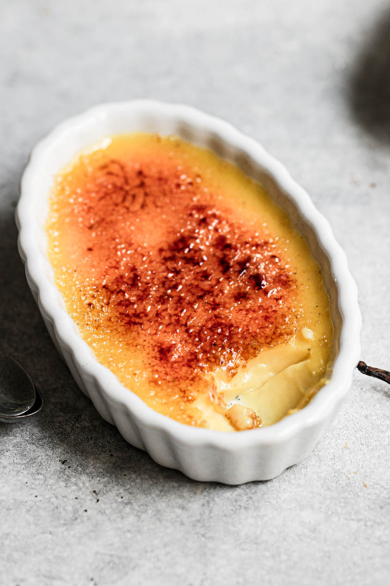 Vanilla bean creme brûlée, baked and torched, in an oval ramekin, with another crème brûlée behind it and 2 small spoons on the side.